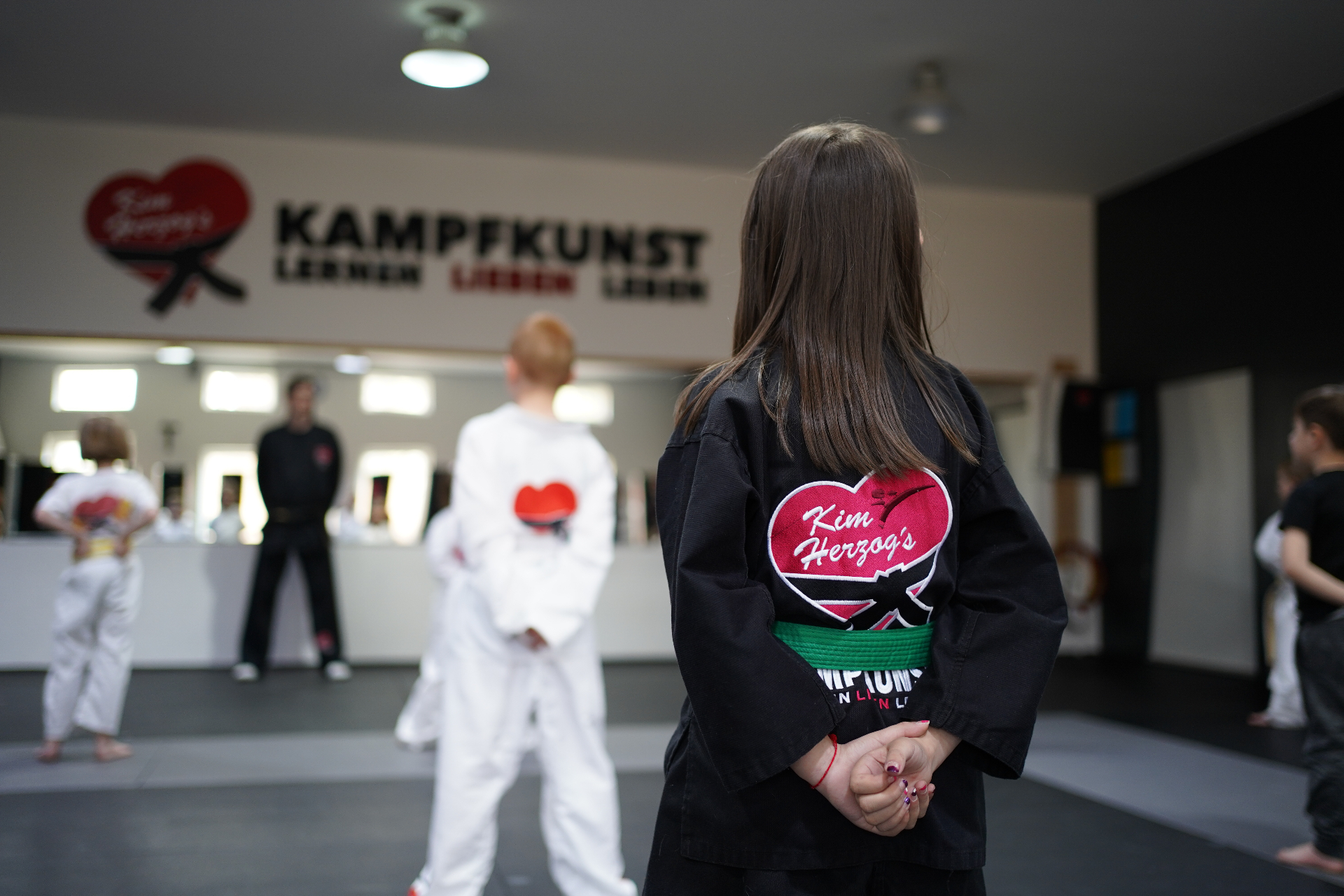 Junges Mädchen in schwarzer Kampfkunst-Uniform mit grünem Gürtel und rotem Herz-Logo im hellen Dojo